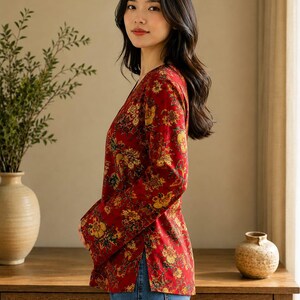 May include: A red floral print top with long sleeves and a V-neck. The top features a pattern of yellow and gold flowers. The person is wearing blue jeans. The background includes a wooden cabinet and a vase with greenery.