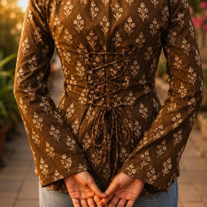 Puede incluir: Top marr&oacute;n de manga larga con estampado floral y espalda con cordones estilo cors&eacute;. El top presenta un cuello alto y mangas acampanadas. El dise&ntilde;o floral es de un tono m&aacute;s claro, creando contraste.