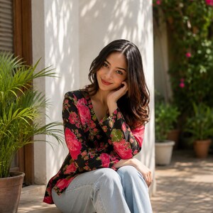 May include: A floral print blazer with a black background and large pink flowers. The long-sleeved jacket is paired with light blue jeans. The model is sitting outdoors near a potted plant and a wall.