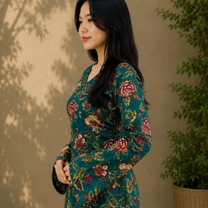 May include: A teal floral print top with long sleeves and a V-neck. The top features a pattern of red and gold flowers and green leaves. The model is wearing blue jeans. The background is a neutral color with shadows.