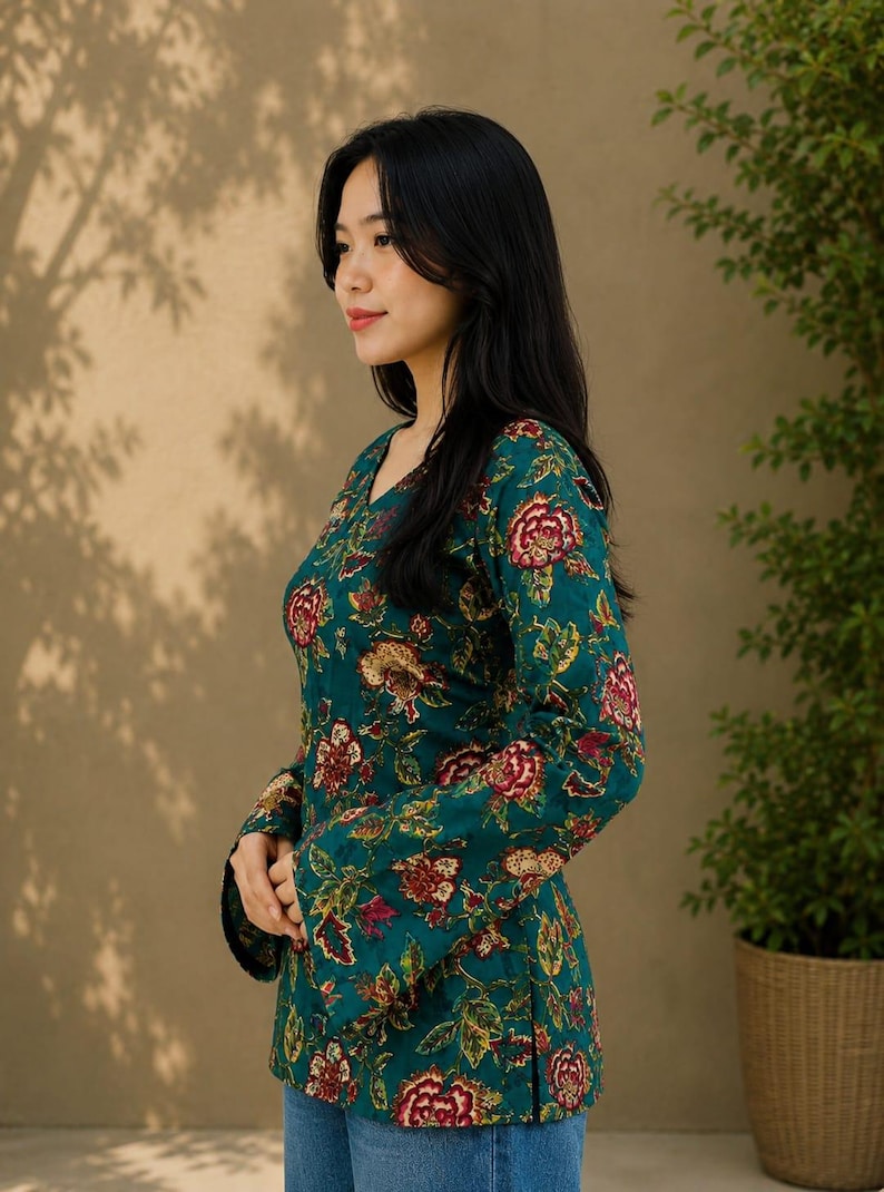 May include: A teal floral print top with long sleeves and a V-neck. The top features a pattern of red and gold flowers and green leaves. The model is wearing blue jeans. The background is a neutral color with shadows.