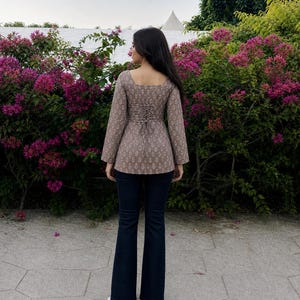 Puede incluir: Una blusa marr&oacute;n estampada con cuello cuadrado y mangas largas, con espalda estilo cors&eacute;. Combinada con vaqueros acampanados azul oscuro y zapatos blancos. El conjunto se presenta sobre un fondo de vibrantes flores rosas y vegetaci&oacute;n.