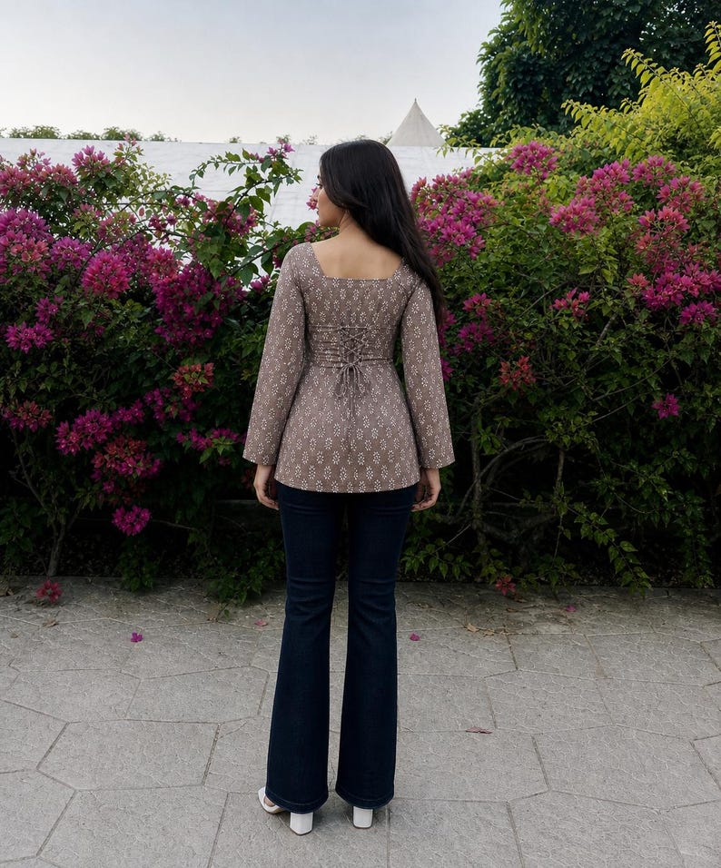 Puede incluir: Una blusa marr&oacute;n estampada con cuello cuadrado y mangas largas, con espalda estilo cors&eacute;. Combinada con vaqueros acampanados azul oscuro y zapatos blancos. El conjunto se presenta sobre un fondo de vibrantes flores rosas y vegetaci&oacute;n.