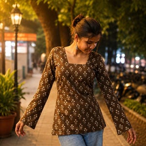 Puede incluir: Una blusa marr&oacute;n de manga larga con cuello cuadrado y estampado floral blanco. La blusa se combina con vaqueros azul claro. La foto est&aacute; tomada al aire libre por la noche, con farolas y &aacute;rboles al fondo.
