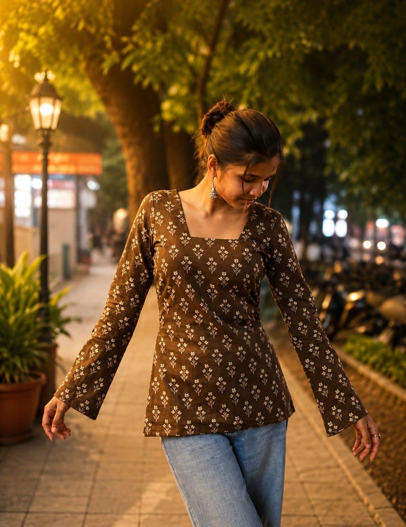 Puede incluir: Una blusa marr&oacute;n de manga larga con cuello cuadrado y estampado floral blanco. La blusa se combina con vaqueros azul claro. La foto est&aacute; tomada al aire libre por la noche, con farolas y &aacute;rboles al fondo.