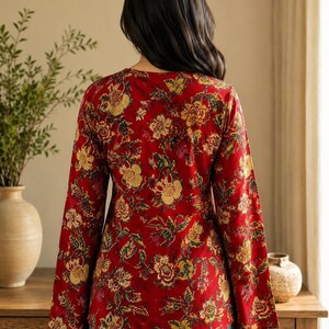 May include: A red floral print top with long sleeves and a V-neck. The top features a vibrant pattern of yellow, brown, and green flowers and leaves. The sleeves are flared at the wrist. The model is wearing blue jeans.