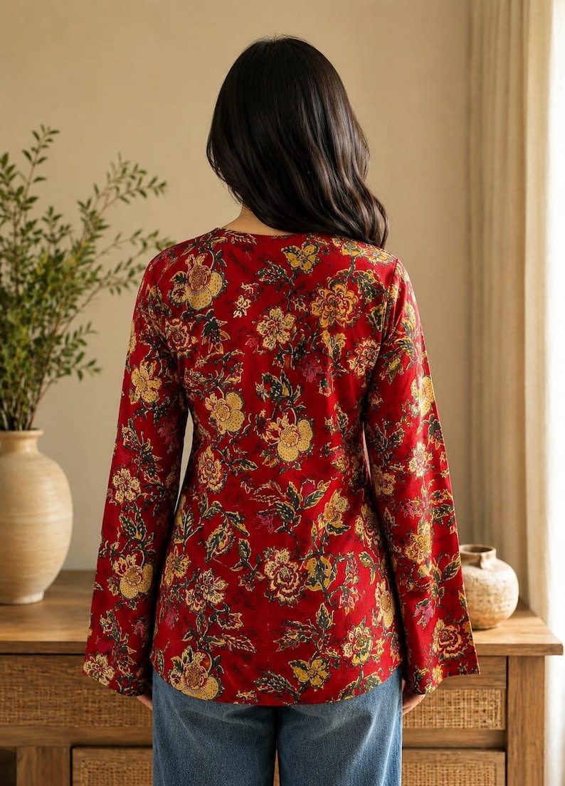 May include: A red floral print top with long sleeves and a V-neck. The top features a vibrant pattern of yellow, brown, and green flowers and leaves. The sleeves are flared at the wrist. The model is wearing blue jeans.