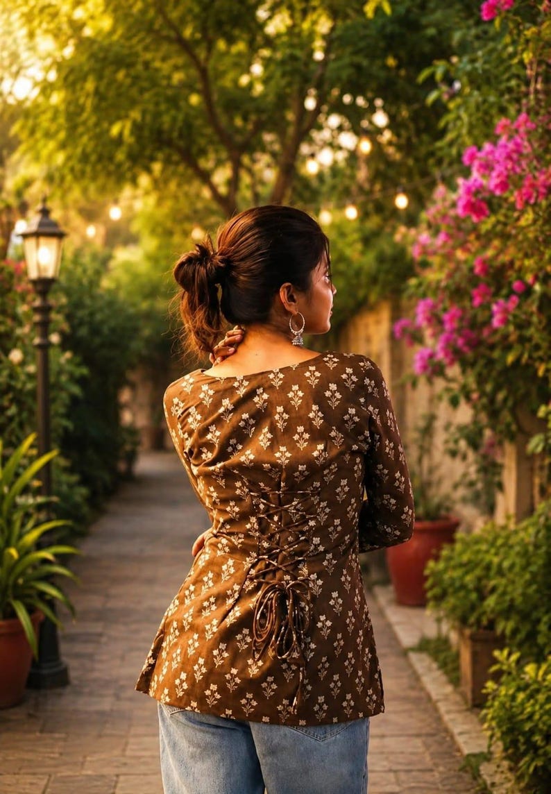 Puede incluir: Una blusa marr&oacute;n con un estampado floral blanco y una espalda con cordones. La blusa de manga larga se lleva con vaqueros azules. El fondo presenta un camino de ladrillos, vegetaci&oacute;n exuberante y flores rosas.