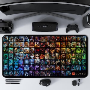 May include: A rectangular gaming mousepad featuring a grid of colorful character portraits from the game DOTA 2. The mousepad is black with the DOTA 2 logo in the bottom right corner. Other gaming accessories are visible.