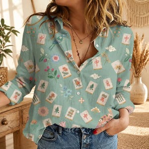 May include: A light green long-sleeve button-down shirt featuring a repeating pattern of mahjong tiles, colorful floral motifs, stylized birds, and clouds. The shirt has a classic collar, a front button placket, and long sleeves with cuffs.