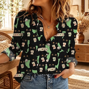 May include: A long-sleeved button-down shirt featuring a repeating pattern of black cats holding mahjong tiles and dice against a light green background. The shirt has a V-neckline, a collar, and cuffs that are rolled up to the mid-forearm.
