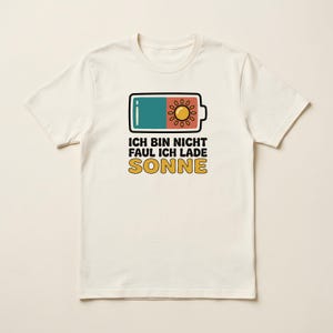 Puede incluir: Una camiseta de manga corta de color crema con un gráfico de un icono de batería que está medio lleno de un color verde azulado y medio lleno de un icono de sol amarillo. Debajo del gráfico está el texto en negro y amarillo: ICH BIN NICHT FAUL ICH LADE SONNE.