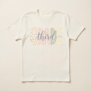 Könnte beinhalten: Ein cremefarbenes Kurzarm-T-Shirt mit dem Wort GRADE in großen, gemusterten Pastellbuchstaben, wobei das Wort third in einer dunkelblauen Schreibschrift über die Mitte der Buchstaben gelegt ist.