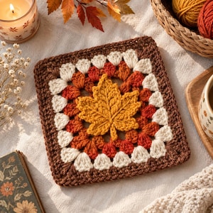 Op de afbeelding: Een vierkant gehaakt granny square met een centraal, gestructureerd geel esdoornbladmotief. Het vierkant is gewerkt in concentrische rondes van crème, oranje en donkerbruin garen, wat zorgt voor een gelaagd, herfstachtig textielontwerp.