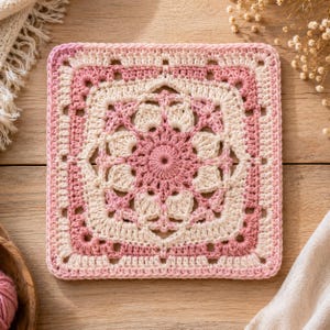 May include: A square crochet motif featuring a central floral pattern in shades of cream and soft pink. The design consists of intricate stitches radiating from the center, framed by a geometric border that transitions between the two colors.