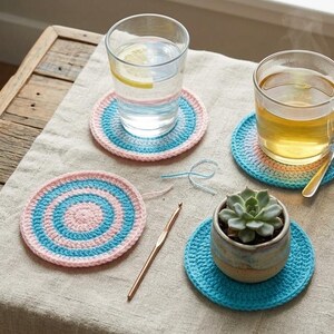 Puede incluir: Un conjunto de posavasos de ganchillo hechos a mano en tonos rosa y azul, dispuestos sobre un mantel de lino neutro. Los posavasos son circulares con anillos concéntricos. Una pequeña suculenta en una maceta de cerámica.