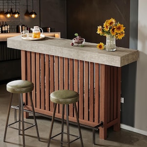 May include: A home bar with a concrete countertop and a wooden slatted front. Two green leather-topped stools with metal legs are in front of the bar. A vase of sunflowers and a bowl of cherries sit on the bar.
