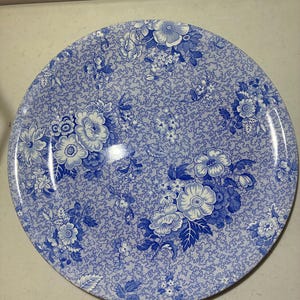 May include: Round ceramic plate with a blue and white floral pattern. The plate features a detailed design of various flowers and foliage against a textured background. The color palette is primarily shades of blue and white.