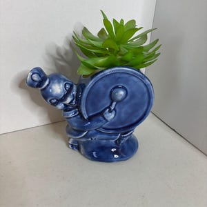 May include: A blue ceramic planter shaped like a drummer playing a drum. The planter has a glossy finish and is filled with a green succulent plant. The drummer figure is detailed with a hat and uniform.