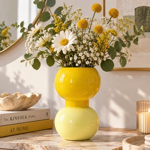 May include: A two-tiered glass vase with a bright yellow top section and a pale yellow bottom section, filled with a bouquet of white daisies, yellow billy balls, small white baby's breath flowers, and green eucalyptus leaves.