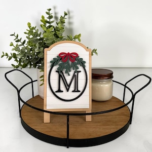 May include: A white wooden sign with horizontal lines features a black circular frame enclosing the letter M. Above the letter is a green mistletoe sprig topped with a red bow. The sign stands on a round wooden tray with a black metal rim.