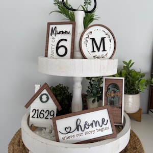 May include: A two-tiered, white wooden tray with decorative signs. The signs include a house number, a welcome sign, a sign that says "home where our story begins", and a sign that says "Party of 6". The tray is decorated with greenery.