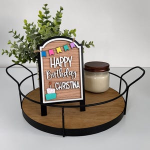 May include: A wooden arched sign with a faux wood grain finish featuring a colorful bunting banner, a white cake with a candle, and the text "HAPPY Birthday CHRISTINA" in raised white lettering, displayed on a round tray with a candle.