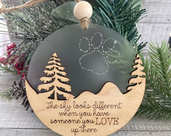 May include: A wooden Christmas ornament with a paw print and two pine trees. The ornament has a frosted glass background and the text "The sky looks different when you have someone you LOVE up there".