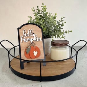 May include: A round wooden tray with a black metal frame and handles, holding a small arched sign with the text "hello Pumpkin" above an orange pumpkin cutout, a green artificial plant in a white pot, and a candle in a glass jar with a lid.