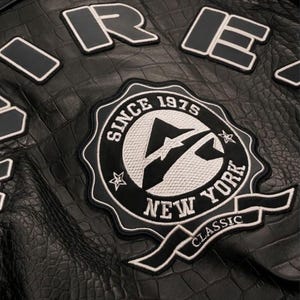 May include: Close-up of a black leather jacket with a crocodile texture. The jacket features patches with white and black lettering, including "ORE" and a circular emblem that reads "SINCE 1975 NEW YORK CLASSIC".