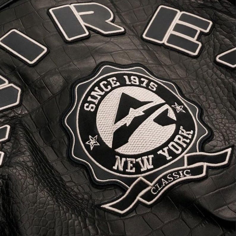 May include: Close-up of a black leather jacket with a crocodile texture. The jacket features patches with white and black lettering, including "ORE" and a circular emblem that reads "SINCE 1975 NEW YORK CLASSIC".