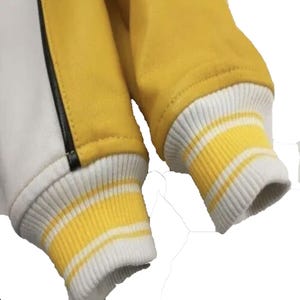 以下が含まれることがあります： イエローの袖と白いボディが特徴のバーシティジャケットのクローズアップ。袖口は白と黄色のストライプ。黒いストライプが白いパネルに沿って走っています。ジャケットは滑らかな素材で作られているようです。