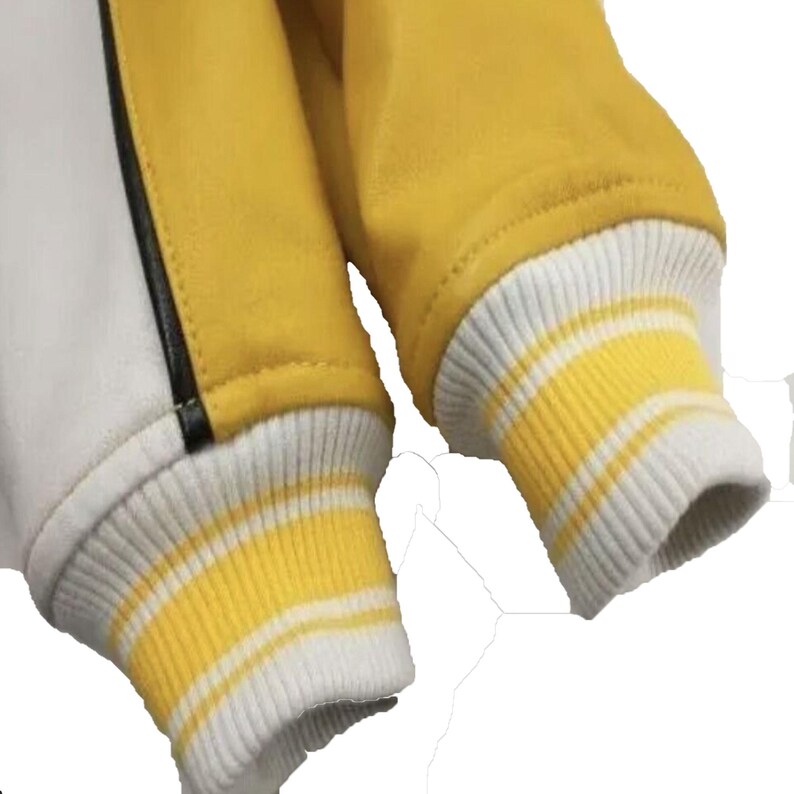 以下が含まれることがあります： イエローの袖と白いボディが特徴のバーシティジャケットのクローズアップ。袖口は白と黄色のストライプ。黒いストライプが白いパネルに沿って走っています。ジャケットは滑らかな素材で作られているようです。