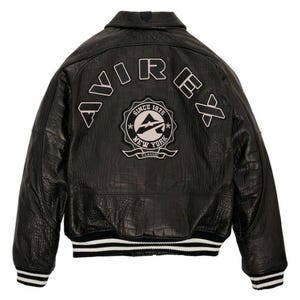 May include: Black leather jacket with a textured finish, featuring the word "AVIREX" across the back in white lettering. A circular patch with "NEW YORK CLASSIC" and "SINCE 1975" is also visible. The jacket has a ribbed hem and cuffs with white stripes.