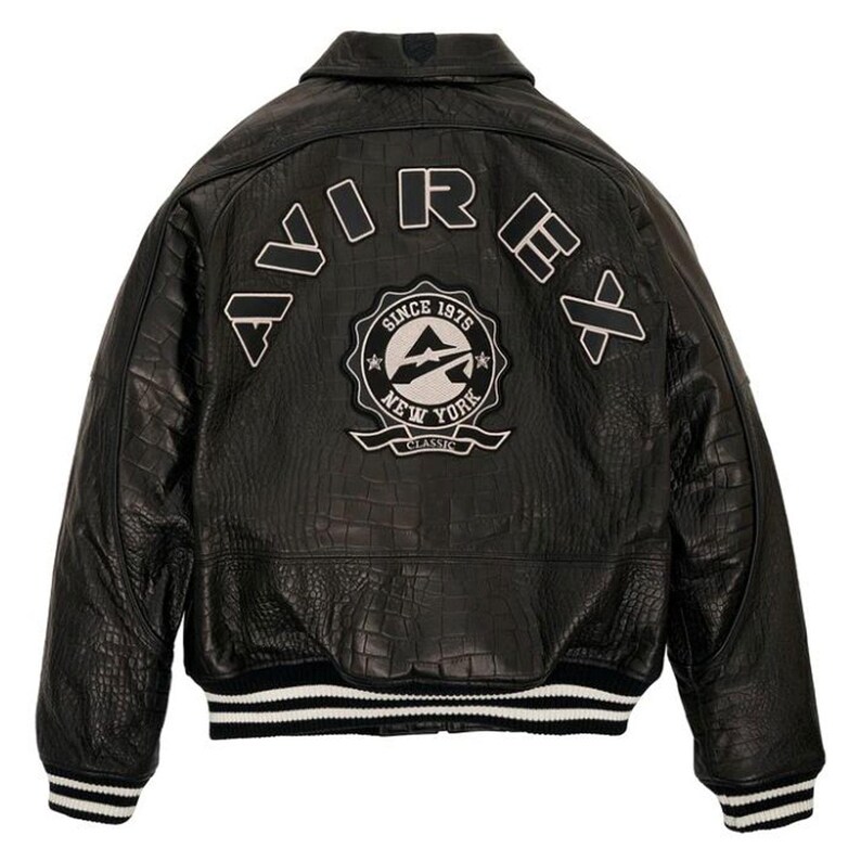 May include: Black leather jacket with a textured finish, featuring the word "AVIREX" across the back in white lettering. A circular patch with "NEW YORK CLASSIC" and "SINCE 1975" is also visible. The jacket has a ribbed hem and cuffs with white stripes.