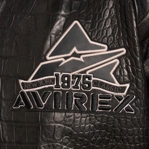 May include: Close-up of a black leather jacket with a textured, crocodile-like pattern. The jacket features a patch with the text "NEW YORK 1975 CLASSIC AVIREX" in white and black. The patch has a triangular design.