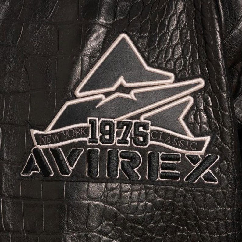 May include: Close-up of a black leather jacket with a textured, crocodile-like pattern. The jacket features a patch with the text "NEW YORK 1975 CLASSIC AVIREX" in white and black. The patch has a triangular design.