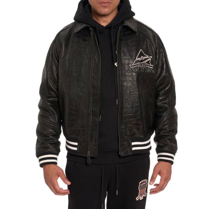 May include: Black leather jacket with a crocodile skin texture. The jacket has a zipper closure, a collar, and white stripes on the cuffs and hem. The Avirex logo is embroidered on the chest.