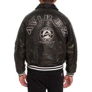 May include: Black leather jacket with a crocodile texture. The back of the jacket features the word "AVIREX" in white lettering and a circular emblem that reads "SINCE 1975 NEW YORK CLASSIC". The jacket has a white and black striped trim.