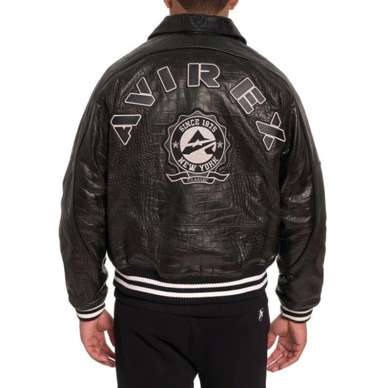 May include: Black leather jacket with a crocodile texture. The back of the jacket features the word "AVIREX" in white lettering and a circular emblem that reads "SINCE 1975 NEW YORK CLASSIC". The jacket has a white and black striped trim.