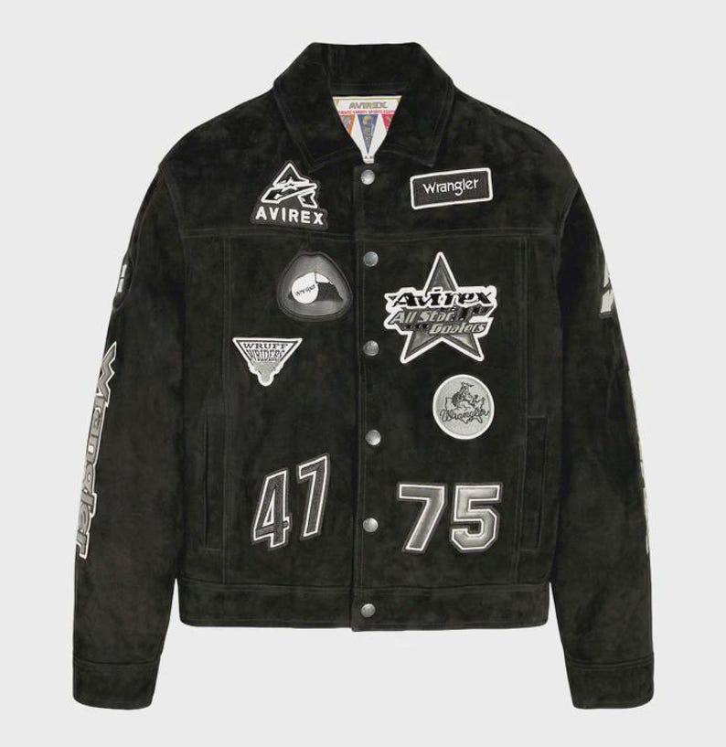 Puede incluir: Una chaqueta de ante negro con cierre de botones, cuello en punta y mangas largas. La parte delantera est&aacute; decorada con varios parches bordados, incluyendo el texto Avirex, Wrangler y los n&uacute;meros 47 y 75 en blanco y gris.