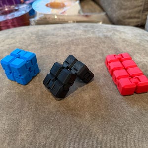 May include: Three fidget toys in the image. A blue cube, a black articulated cube, and a red articulated rectangle. The toys are made of plastic and are designed to be manipulated. The background is a neutral color.