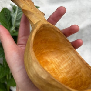 May include: A hand-carved wooden kuksa cup featuring a smooth, light-brown finish with visible wood grain patterns. The cup has a deep, rounded bowl and an integrated handle with carved detailing on the top surface, showcasing a natural texture.