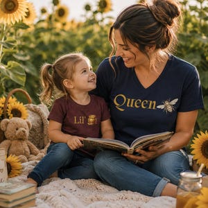 Puede incluir: Una camiseta azul marino con cuello en V con la palabra Queen y un gráfico de abeja en dorado, y una camiseta granate con el texto Lil' y un gráfico de tarro de miel dorado. Ambas camisetas tienen mangas cortas y cuello redondo.