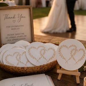 May include: A woven basket filled with round white coasters featuring a gold heart-shaped wheat stalk design, accompanied by a wooden sign that reads Thank You for celebrating with us, and an open guest book with a gold pen.