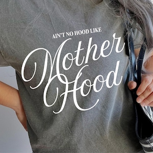 May include: Gray t-shirt with the text "AIN'T NO HOOD LIKE Mother Hood" in white script. The shirt is short-sleeved and appears to be made of a soft, worn material. A black and white striped tote bag is visible.