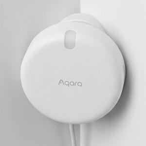 Puede incluir: Un dispositivo doméstico inteligente Aqara blanco montado en una esquina de la pared. El dispositivo tiene una forma redondeada con una pequeña abertura ovalada y el nombre de la marca impreso en la parte delantera. Dos cables blancos se extienden desde la parte inferior.