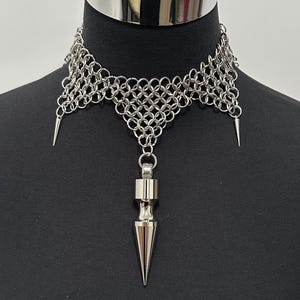 Puede incluir: Un collar tipo gargantilla de cota de malla plateada que presenta una serie de anillos metálicos entrelazados. El diseño incluye dos pequeñas púas que apuntan hacia abajo en los lados y un colgante central más grande de metal pulido.