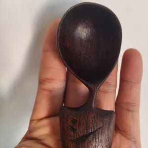 May include: A handcrafted wooden spoon with a dark brown finish. The spoon has a rounded bowl and a unique handle design, with carved details on the base. The spoon is held in a hand.