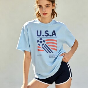 Op de afbeelding: Lichtblauw T-shirt met "U.S.A" in marineblauw, een voetbal en een vlagontwerp. Het shirt heeft de tekst "Est.1913" en "United States of America Soccer Club". Het model draagt marineblauwe shorts met witte bies.